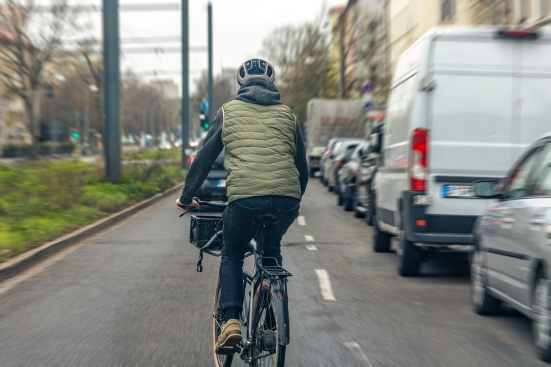 Ciclista com capacete pedalando em via urbana compartilhada com carros e outros veículos.