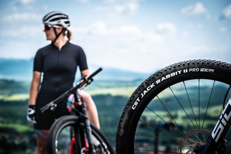 Detalhe do pneu CST Jack Rabbit em uma mountain bike, com ciclista ao fundo em ambiente externo.